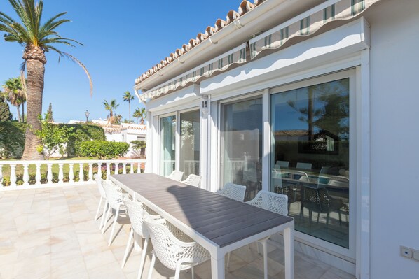 A patio with a wooden table and white chairs, a glass door, and a palm tree. - Villa Marbesa Beach (Marbella)