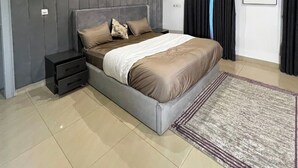 A modern bedroom with a large bed, a nightstand, a wall-mounted panel, a framed picture, and a patterned rug. - JOEY HOMES  (Abuja)