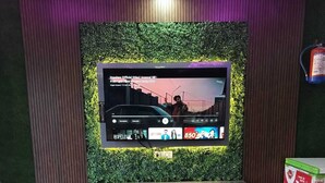 A restaurant interior with a large flat-screen TV mounted on a green wall, wooden tables and chairs, and a branded ice cream dispenser.