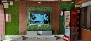 Un interior moderno con un televisor montado que muestra una vista de la calle, una máquina expendedora de Coca-Cola y un panel de madera con follaje verde.