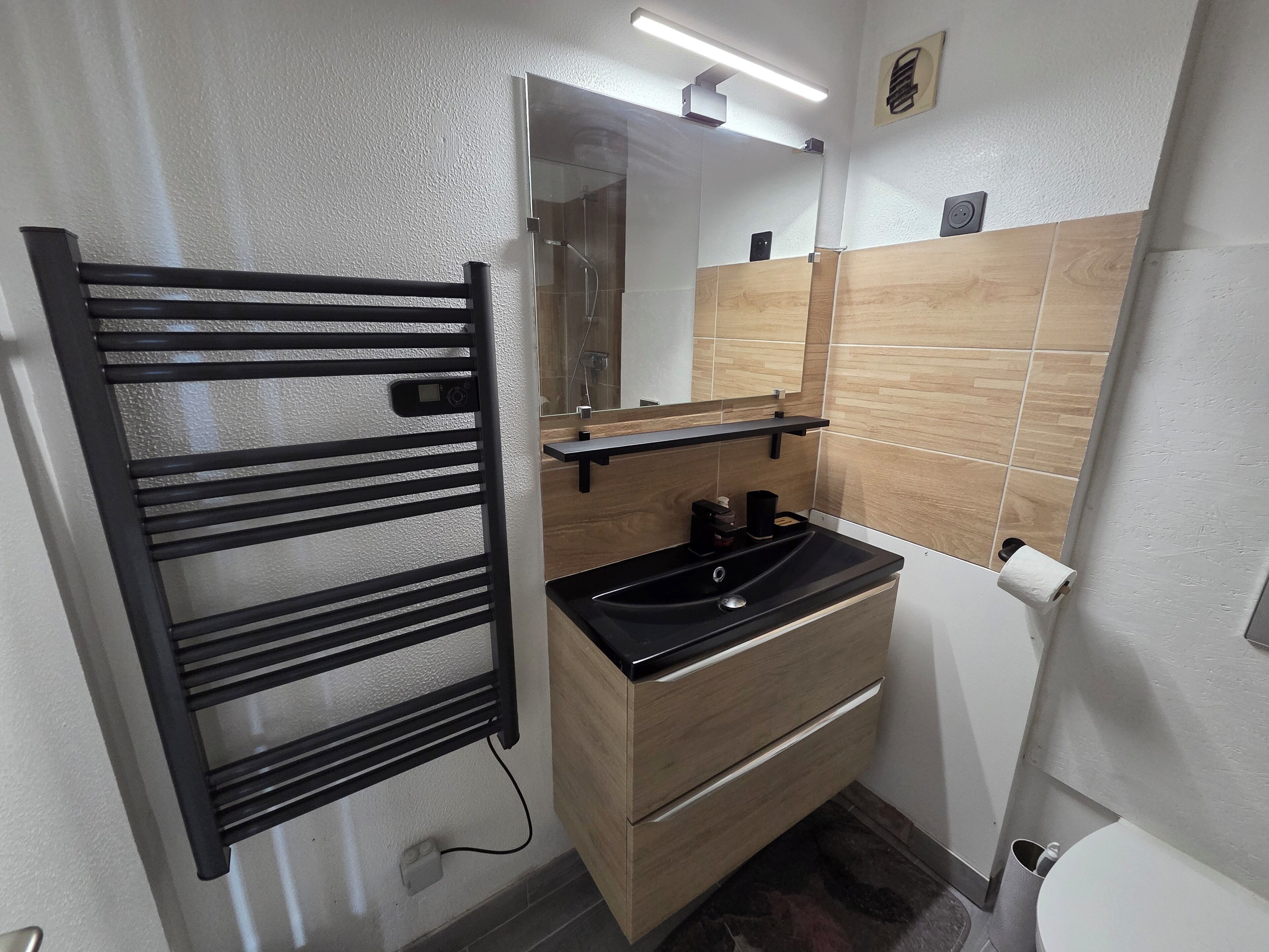 Une salle de bain moderne avec un porte-serviettes noir, un lavabo surmonté d’un miroir et des toilettes.