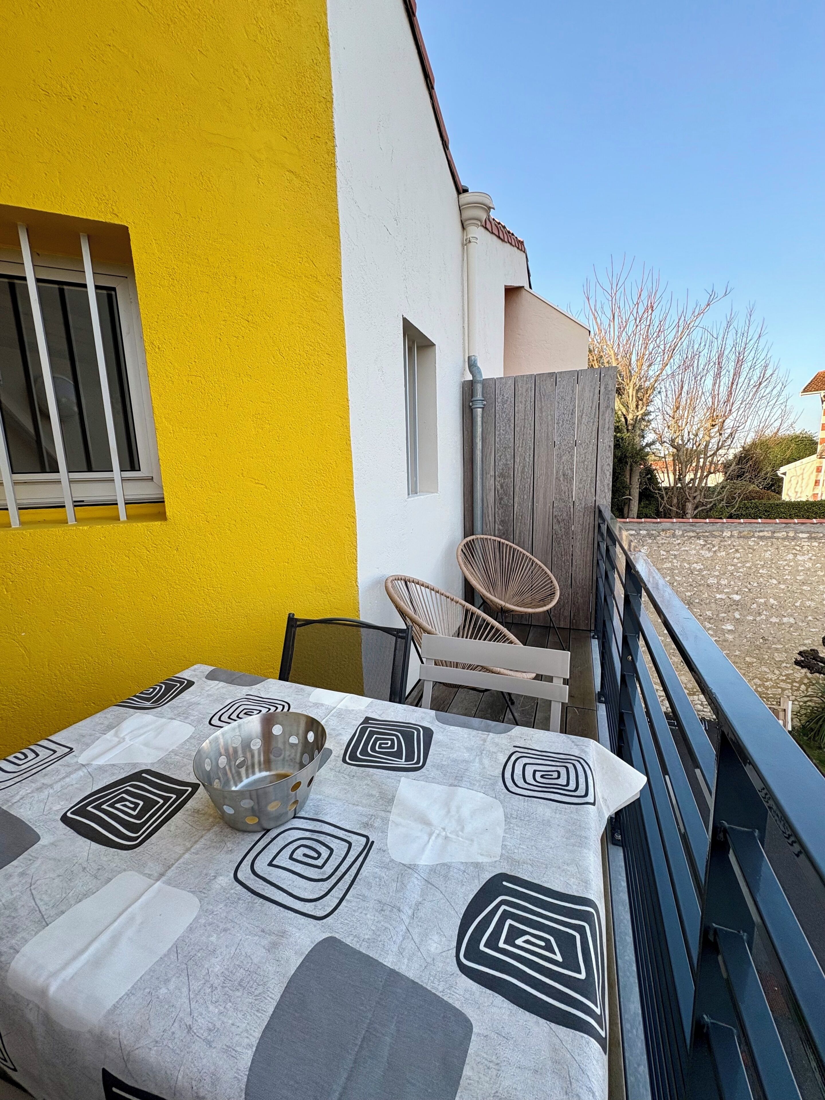 Ein Tisch mit einem gemusterten Tischtuch, zwei Korbstühle und eine Metallschüssel auf einem Balkon mit einer gelben Wand und einem Holenzaun.