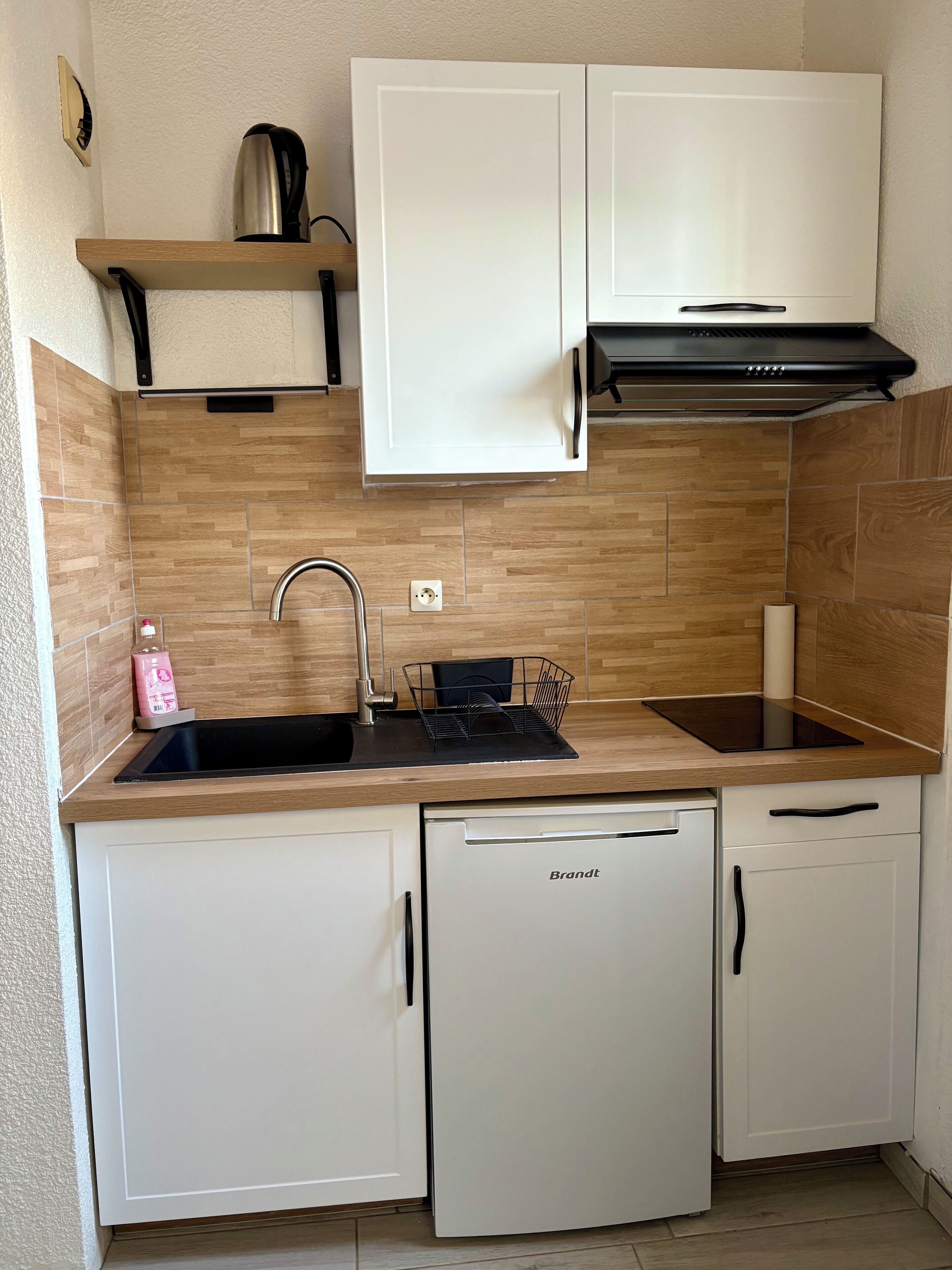 A compact kitchen with white cabinets, a built-in dishwasher, and a stainless steel kettle on a shelf.
