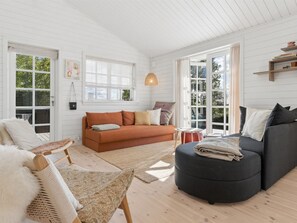 Ein geräumiges Wohnzimmer mit einer Couch, einem Hocker und einem Sessel. Große Fenster und eine Decke mit Holzbalken.
