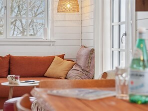 Un cuarto acogedor con una mesa de madera, un sofá con cojines, una ventana con vista a árboles y una lámpara colgante.