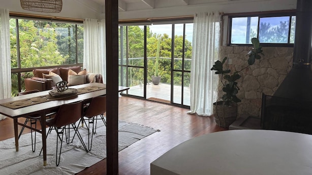 A spacious room with a dining area, a fireplace, and large windows offering a view of greenery. - Surrounded by Nature With Panoramic Mountain Views (Lower Beechmont)