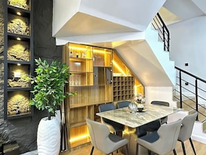 A modern dining area with a marble table, grey chairs, a potted plant, and a staircase. - Yusuf Baba Shortlets (Abuja)