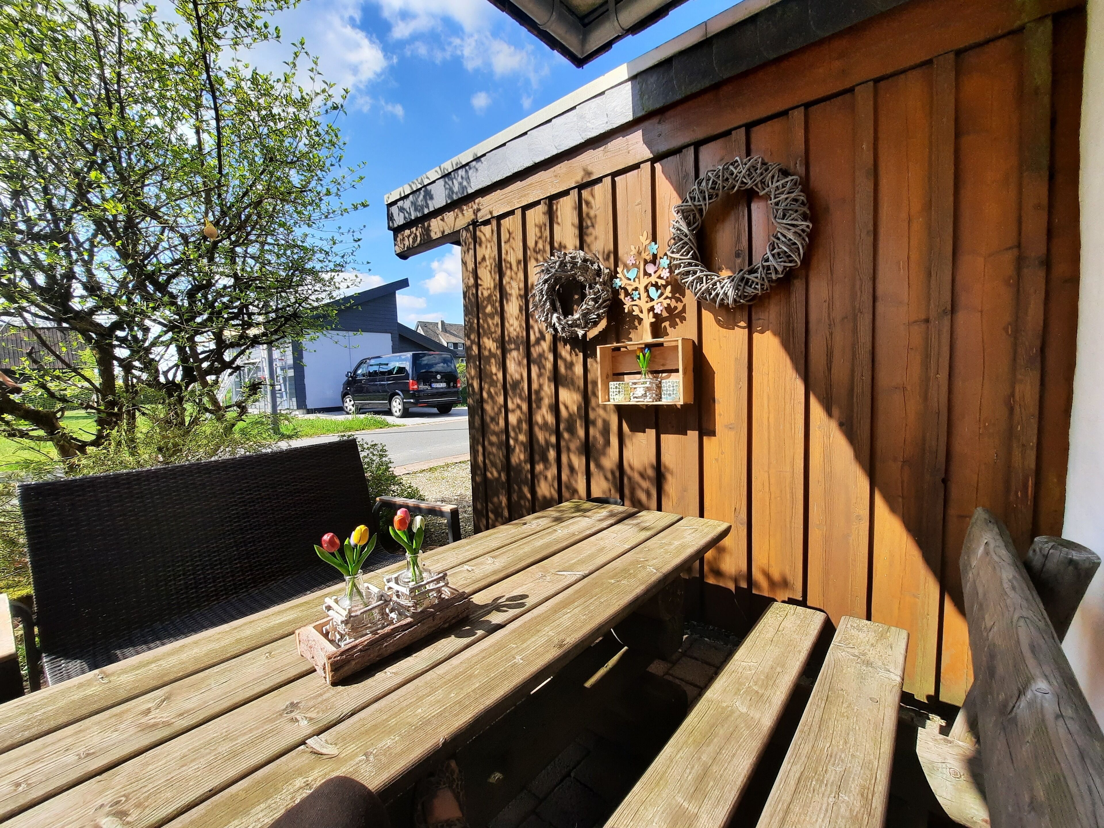 Un espace de détente extérieur en bois, comprenant un banc et une table, décoré d’une guirlande et d’une petite plante.