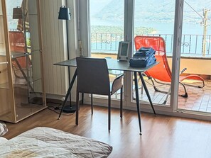 A room with a bed, a table with a lamp, a chair, and a view of a lake and mountains through large windows. - Casa Giovanna Ronco sopra Ascona by Interhome (Ronco sopra Ascona)