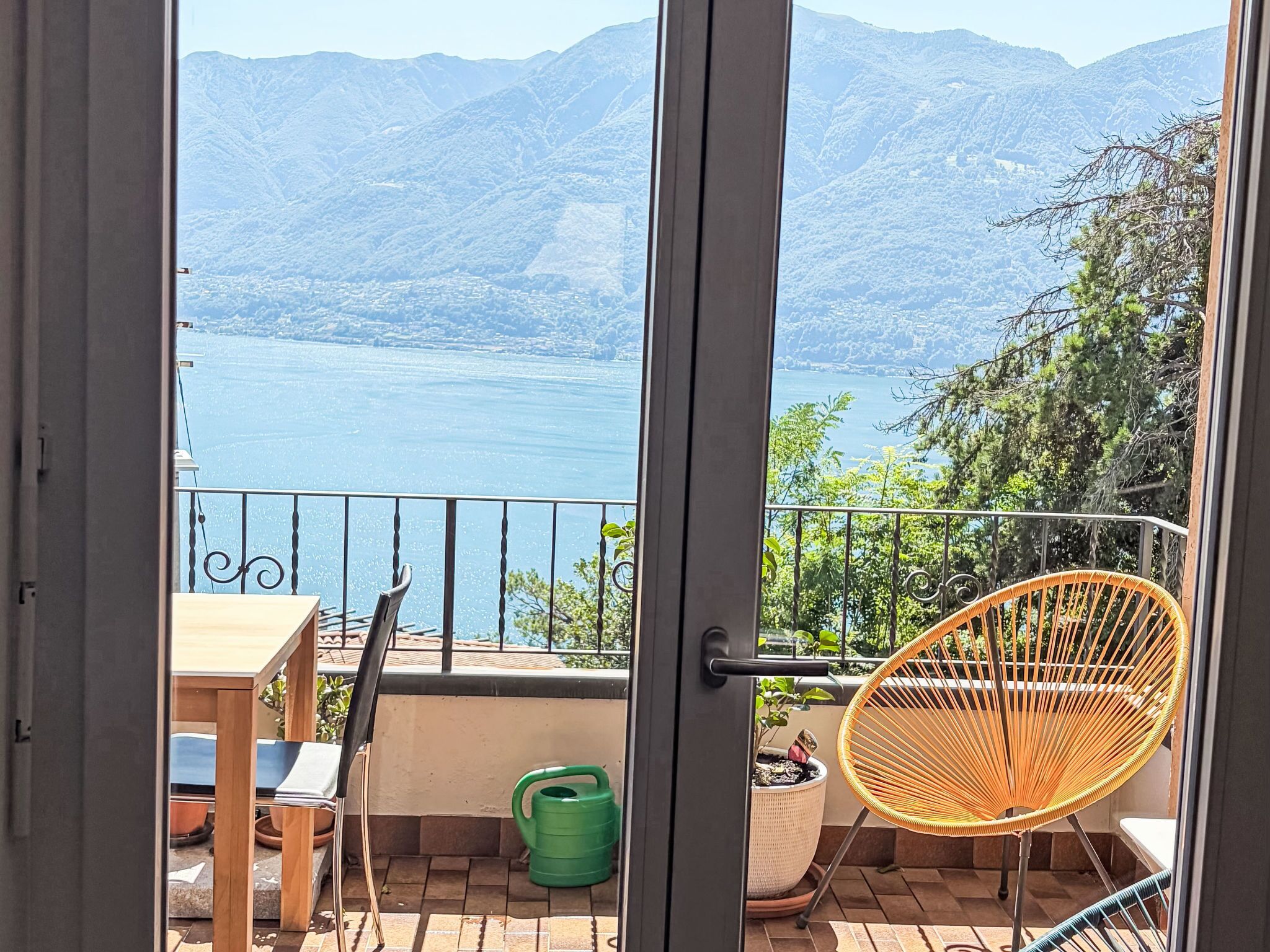 A balcony with a view of a lake and mountains, a wicker chair, a table, and a potted plant.