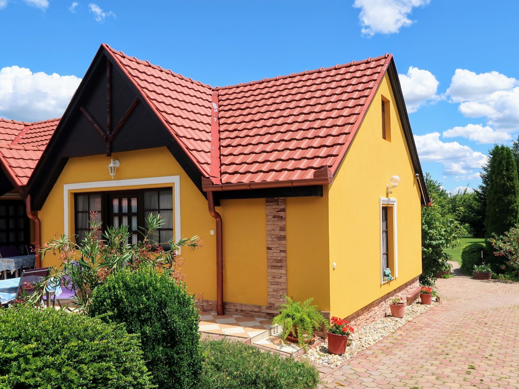 Un bâtiment jaune avec un toit recouvert de tuiles rouges, un mur en pierre servant d’élément décoratif et un petit jardin agrémenté de plantes en pot.