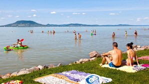 Una playa con gente nadando y haciendo kayak, un área con césped y toallas, y una cadena montañosa distante.