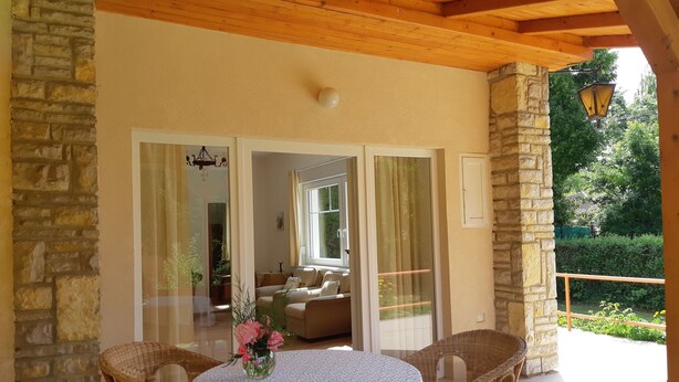 A covered patio area with a round table and wicker chairs, a stone wall, and a view of a garden.