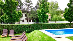 Une piscine dotée d’une terrasse en bois et de chaises longues, entourée d’une pelouse et de haies bien entretenues.