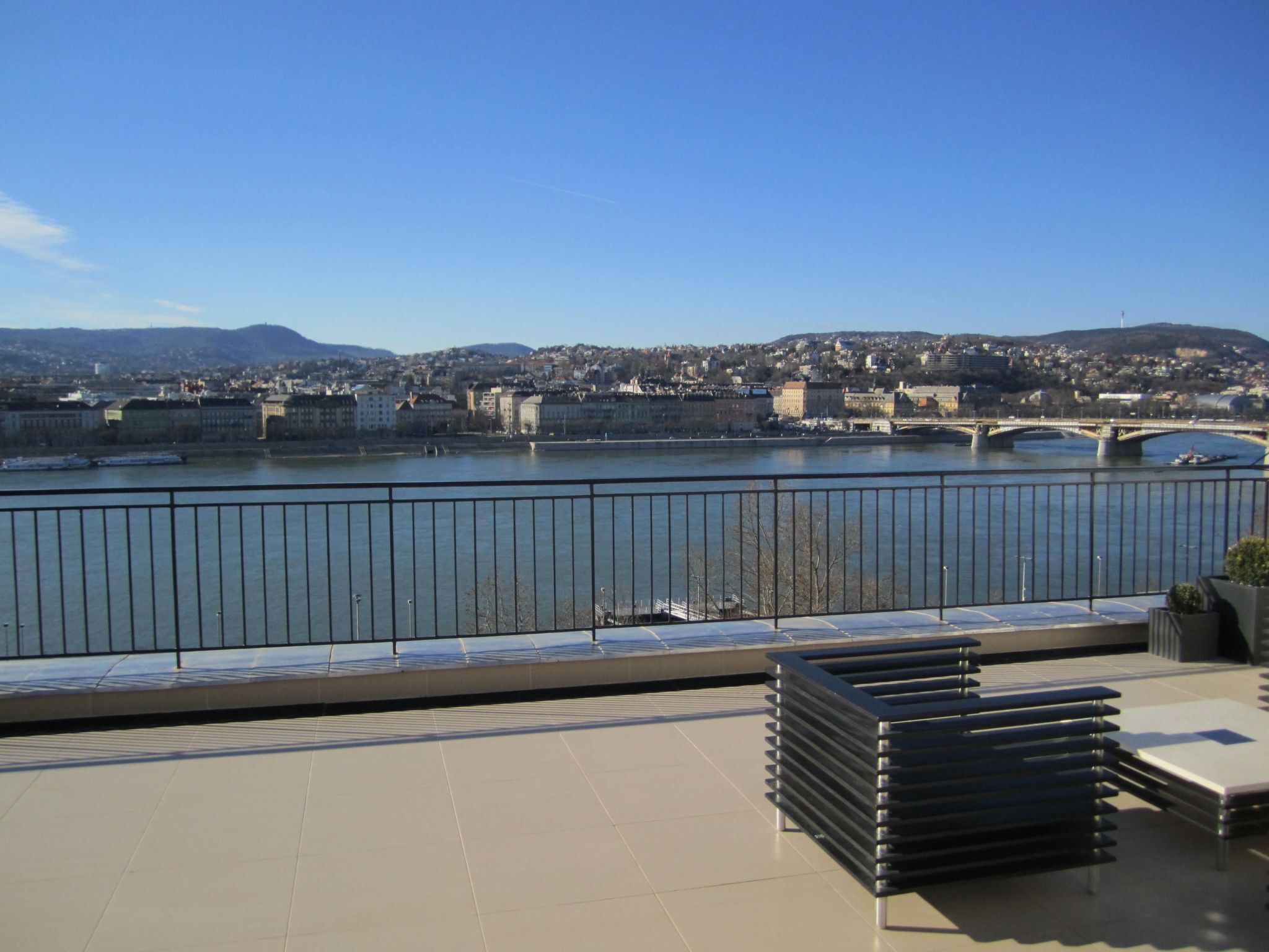 A terrace with a view of a river and a bridge, featuring a railing and a stack of chairs.