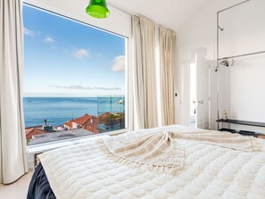 Ein Schlafzimmer mit großem Fenster, einem Bett mit weißer Decke und Blick aufs Meer.