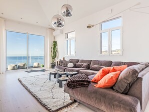Ein modernes Wohnzimmer mit einem großen grauen Sofa, einem weißen Teppich und Meerblick durch große Fenster.