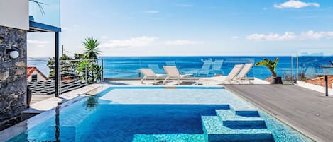 Uma piscina com água azul cristalina, rodeada por espreguiçadeiras e vista para o oceano.