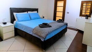 A neatly made bed with blue and gray bedding, a bedside table with a lamp, and a door with a window. - Dimora Mira - Downtown Bari (Bari)