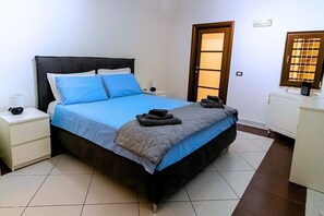 A neatly made bed with blue and gray bedding, a bedside table with a lamp, and a door with a window. - Dimora Mira - Downtown Bari (Bari)