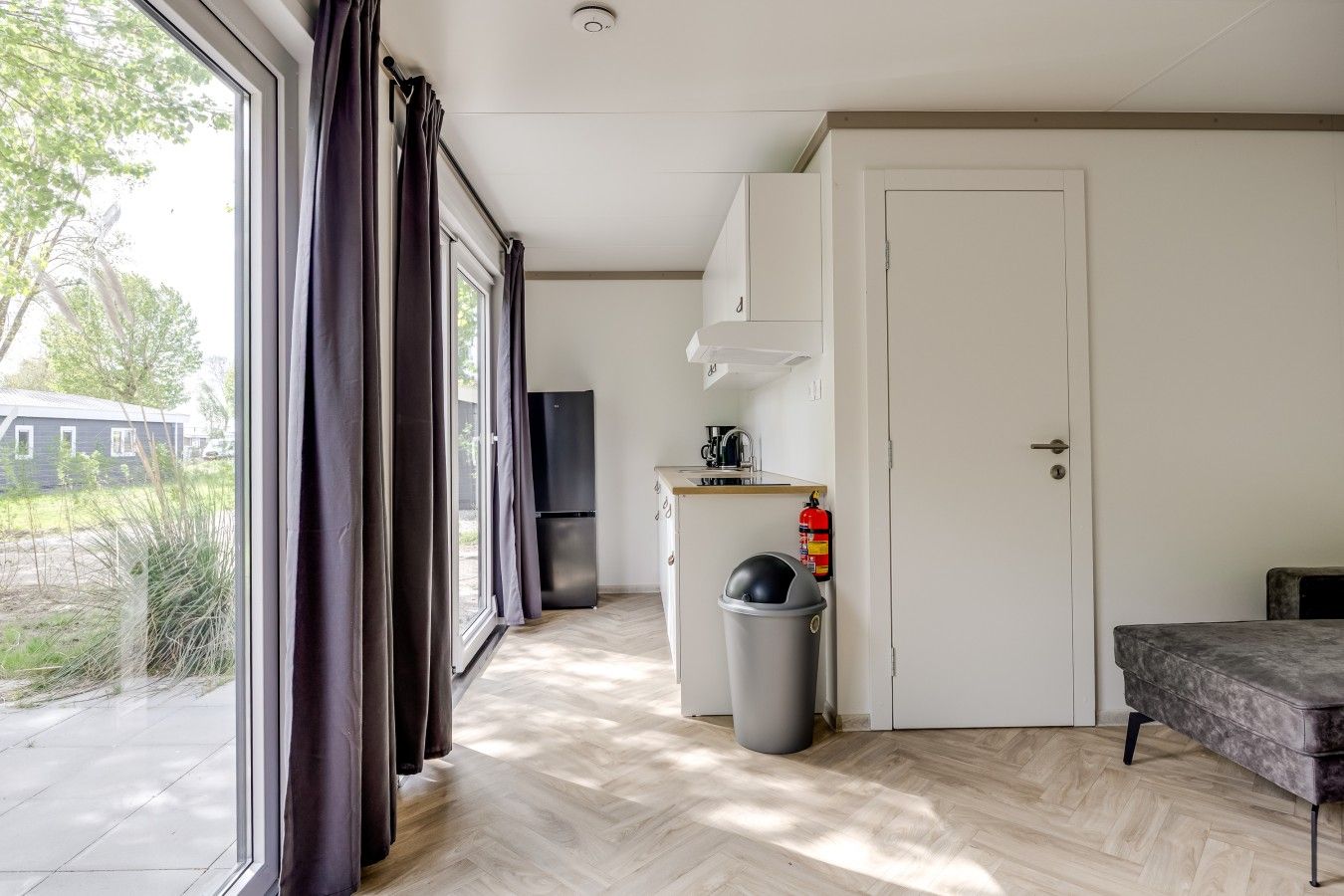 A room with a large window, a door, a kitchen area with a refrigerator, a trash bin, and a small table.