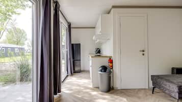 A room with a large window, a door, a kitchen area with a refrigerator, a trash bin, and a small table.