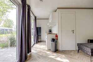 Ein Zimmer mit großem Fenster, einer Tür, einer Küchenzeile inklusive Kühlschrank, einem Mülleimer und einem kleinen Tisch.
