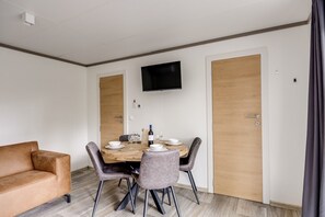 A modern room with a wooden dining table, four chairs, a sofa, a TV mounted on the wall, and two wooden doors.