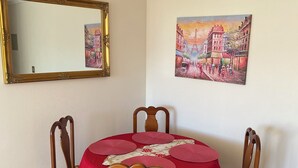 Zona pranzo con un tavolo rotondo ricoperto da una tovaglia rossa, circondato da sedie di legno con cuscini gialli. Un grande specchio è appeso al muro sopra il tavolo, e un quadro raffigurante una scena di strada è esposto sulla parete adiacente.