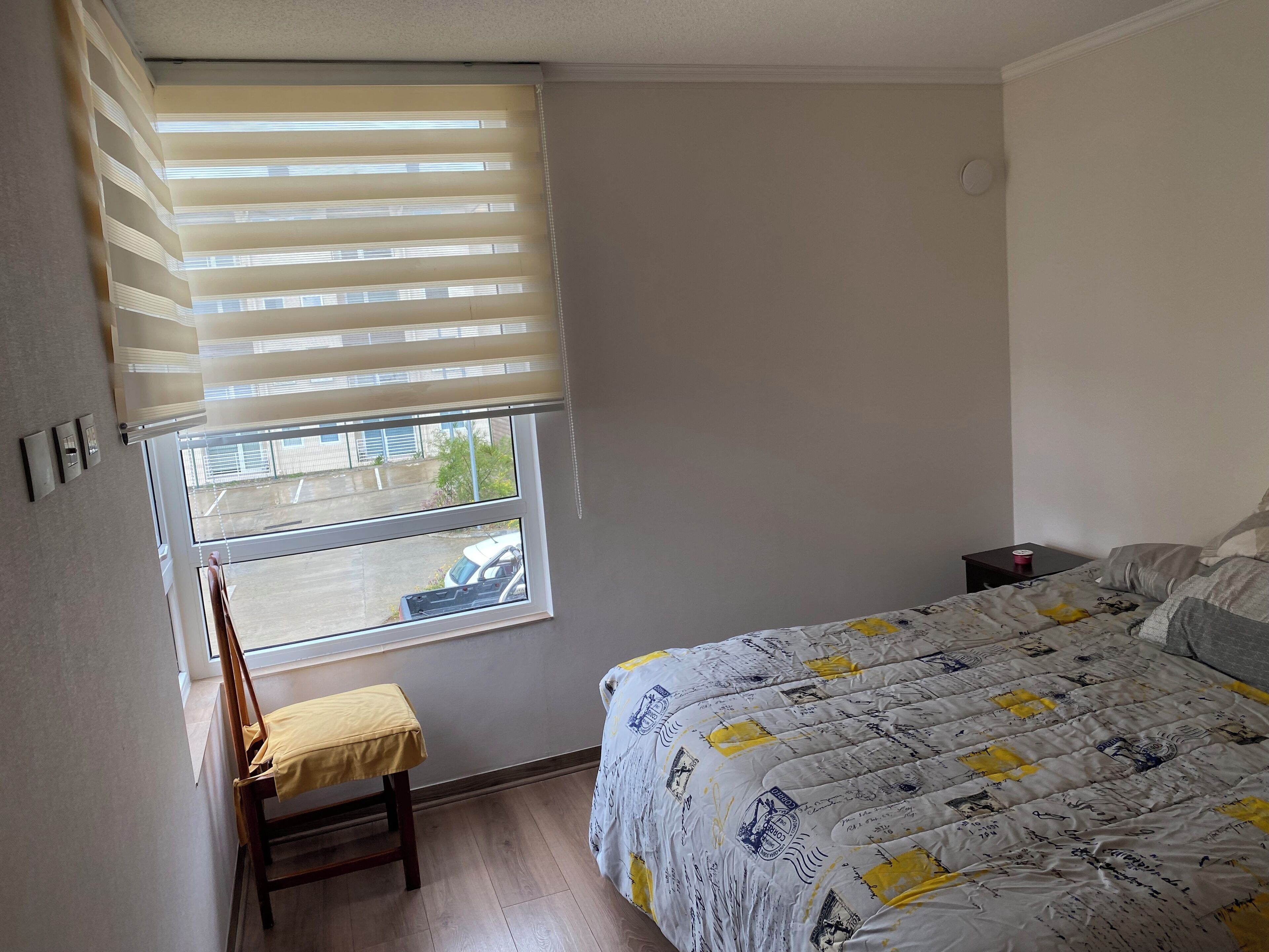 Een slaapkamer met een bed, een stoel, een raam met jaloezieën en een nachtkastje.