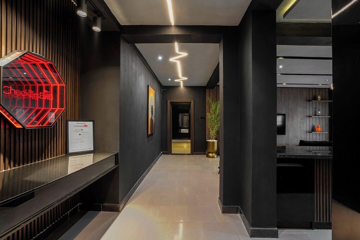 Un hall d’hôtel moderne avec un comptoir d’accueil, un panneau lumineux au néon et un couloir menant à une chambre.