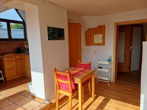 A compact kitchen with wooden cabinets, a dining table with chairs, a small white cabinet, and a door leading to another room.
