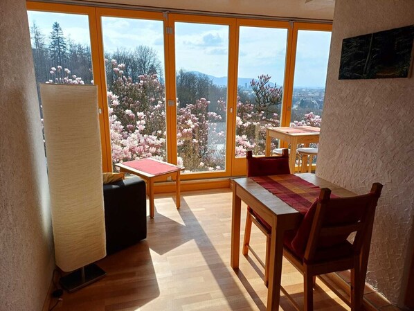 A room with a wooden table and chairs, a small table with a red table runner, and large windows with a view of blooming trees.