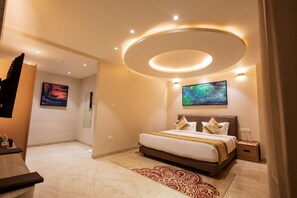 A modern hotel room with a large bed, a flat-screen TV, a ceiling light fixture, and a painting on the wall. - Saaz Hotel - Near Indore International Airport (Indore)