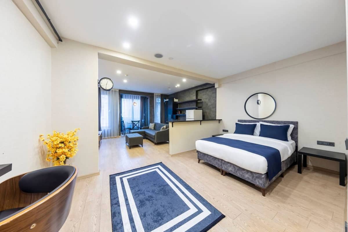 A modern hotel room with a large bed, a desk, a chair, and a small kitchenette.