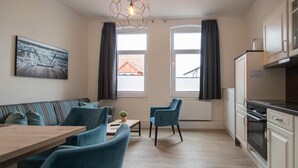 A modern kitchen with white cabinets, a built-in oven, and a countertop. There is a dining area with a wooden table and blue chairs. - Villa Sofia on Borkum - Stylish vacation apartment in a central location (Borkum)