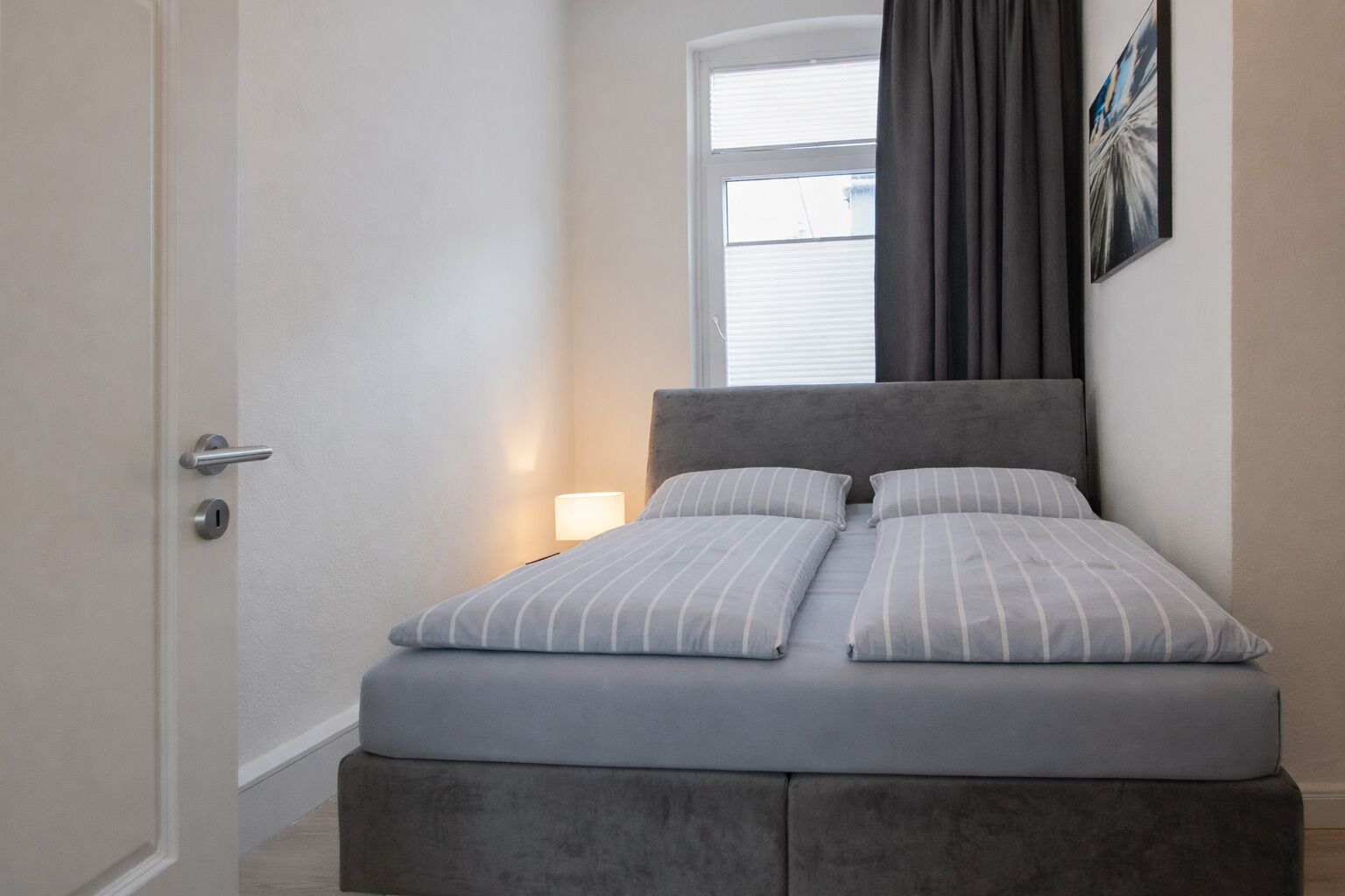 Een slaapkamer met een bed, een nachtkastje, een raam met gordijnen en een schilderij aan de muur.