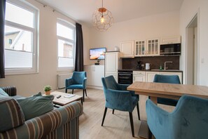 Een gezellig, compact leefruimte met een kitchenette, een bank, twee stoelen, een houten eettafel en een hanglamp.