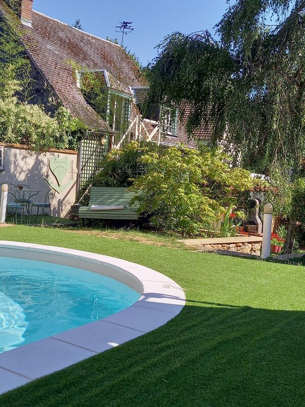 Une piscine entourée d’une pelouse bien entretenue, une maison en briques avec un toit en pente et un escalier en bois menant à une terrasse.