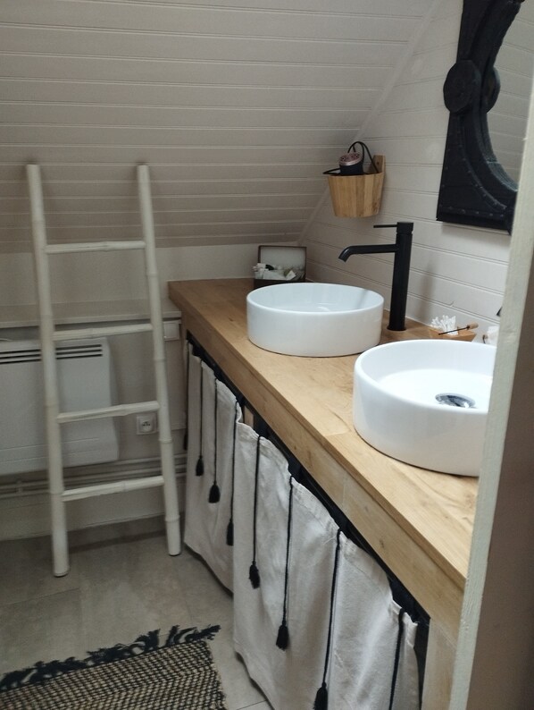A small bathroom with two white sinks, a wooden vanity, a white towel rack, and a black towel hanging.