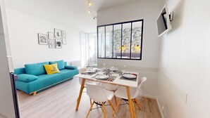 A modern living room with a teal sofa, a dining table set for four, and a large window with a view of trees. - Le Grain de Sel (Saint-Hilaire-de-Riez)