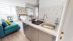 A modern kitchen with a sink, a dish rack, and a dining area with a blue sofa. - Le Grain de Sel (Saint-Hilaire-de-Riez)
