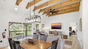 Uma ampla sala de jantar com mesa e cadeiras de madeira, ventilador de teto e uma grande janela com vista para o exterior.