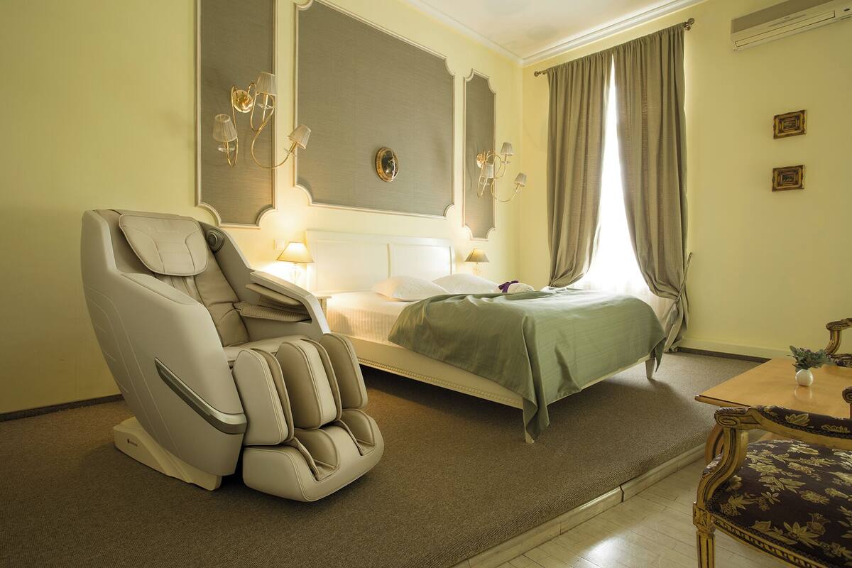 Una habitación de hotel con una silla de masajes, una cama con colcha verde, una mesita de noche con lámpara y una pequeña mesa con un florero.