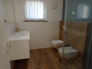 Een moderne badkamer met een wit wastafelmeubel, toilet en een bidet, houten vloeren en een raam met dunne gordijnen.