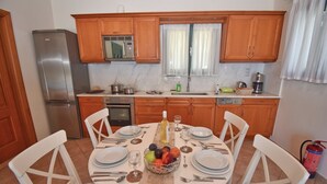 A kitchen with wooden cabinets, a stainless steel refrigerator, a built-in oven, and a dining table set for four with white chairs. - Villa Pasiphae (Corfu)
