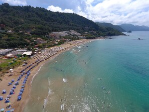 Ein Strand mit Reihen von Sonnenschirmen und Liegestühlen, ein sandiger Uferstreifen und Gebäude im Hintergrund.