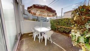 A patio with a white table and chairs, a brown and white umbrella, and a hedge. - Studio des Nénuphars - Beach 500m away & terrace (Saint-Hilaire-de-Riez)
