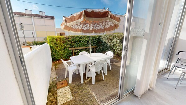 A patio area with a white table and chairs, a brown and white umbrella, and a glass door leading to an indoor space. - Studio des Nénuphars - Beach 500m away & terrace (Saint-Hilaire-de-Riez)