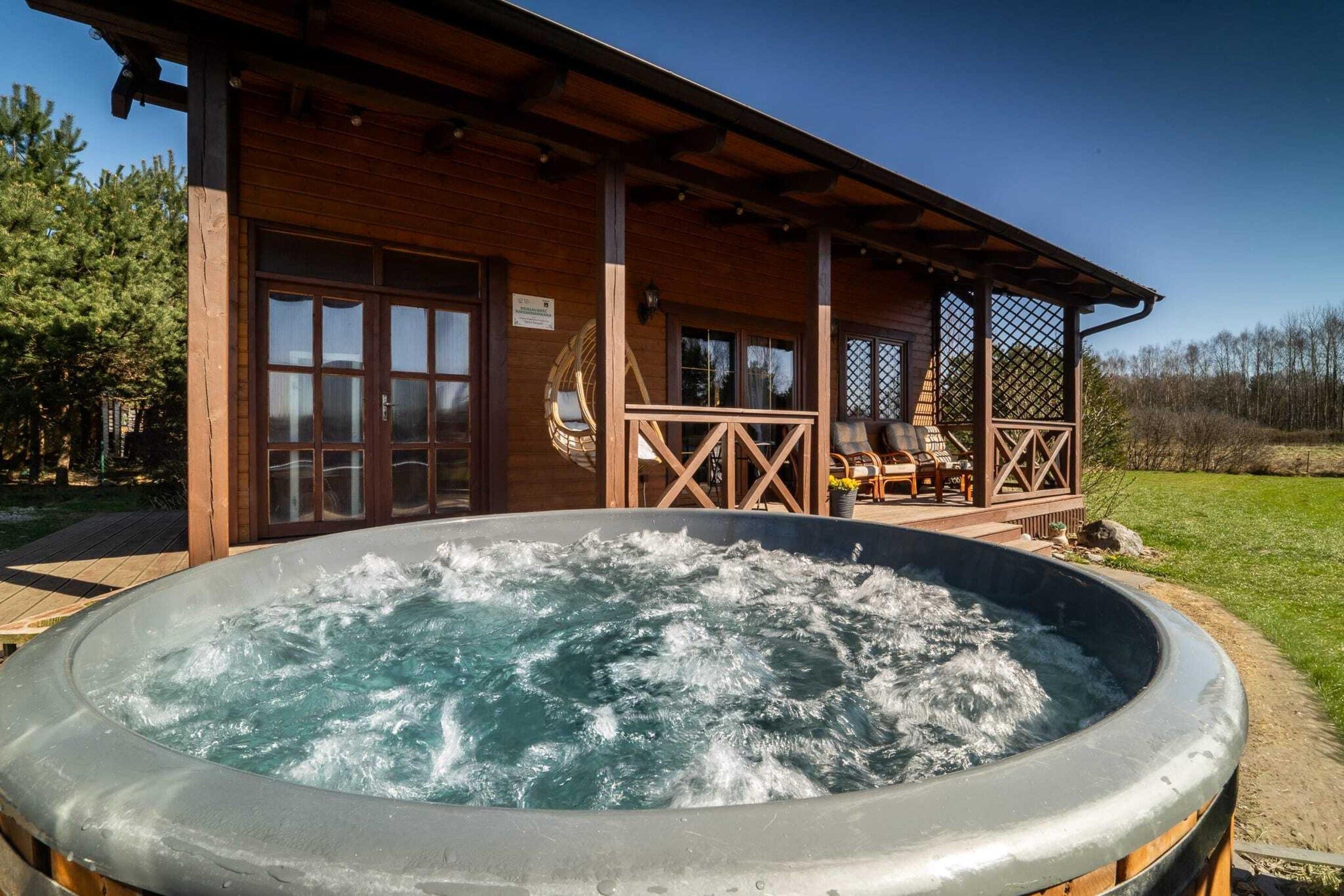 Una cabaña de madera con un jacuzzi en la parte delantera, un porche cubierto con asientos y un cielo despejado.